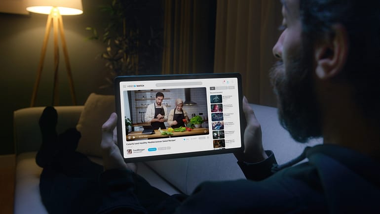 A person watches an instructional cooking video on a tablet while sitting in a dimly lit room.