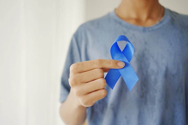 Person wearing a blue shirt holding a blue awareness ribbon between their fingers.