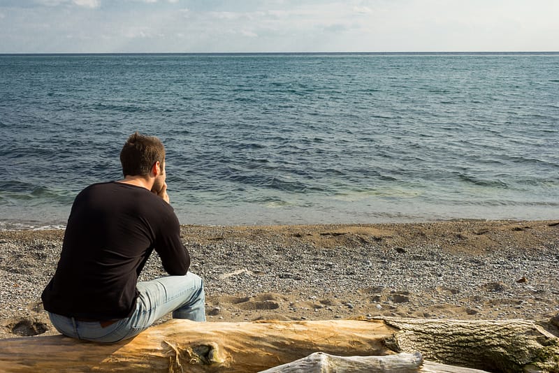 A person sitting alone on a driftwood log at a rocky shoreline, gazing out at calm open water.