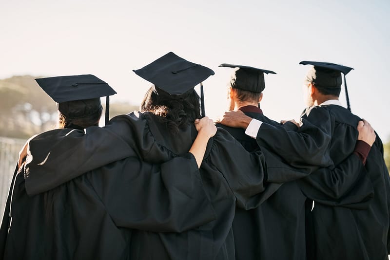 Four graduates in caps and gowns standing arm in arm, facing away from the camera on a bright day.